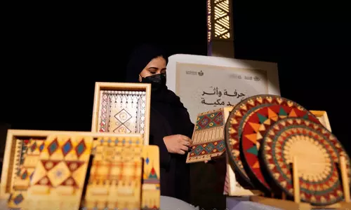 A woman in traditional Saudi attire stands behind a display of handmade crafts and decorative items at a cultural stall during the Makkah Winter Festival, photographed at night.