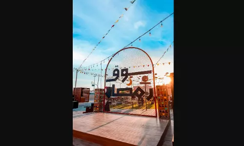 A festive outdoor setting for a Ramadan Souq featuring a wooden stage in the foreground. A large arched wire-mesh sign displays the Arabic text سوق رمضان (Ramadan Market) in bold black lettering, accented with a small yellow crescent moon. The scene is decorated with hanging string lights and colorful pennant banners against a bright blue sky at sunset, with a modern urban backdrop.
