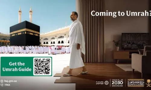 A promotional graphic from the Saudi Ministry of Hajj and Umrah titled Coming to Umrah?. The image is split into two halves: on the left, a vibrant outdoor view of the Kaaba in Makkah surrounded by pilgrims; on the right, a man in white Ihram clothing stands in a modern living room looking toward the Kaaba. A large green box in the corner features a QR code with the text Get the Umrah Guide, and the bottom includes official logos for Saudi Vision 2030 and the Ministry of Hajj and Umrah.