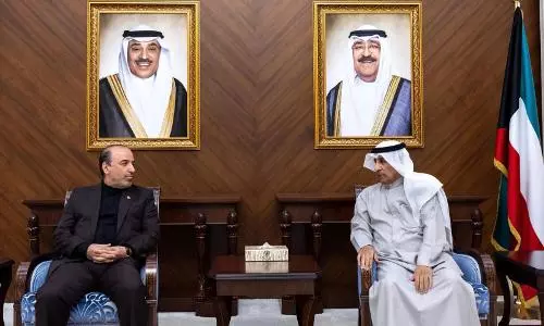 Two officials seated in a formal meeting room beneath portraits of Kuwaiti leaders, with the Kuwaiti flag displayed on the right.
