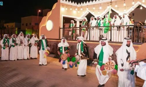 Men in traditional Saudi attire perform a folkloric dance with drums and swords during an evening Eid celebration, as officials stand on a decorated stage behind them.