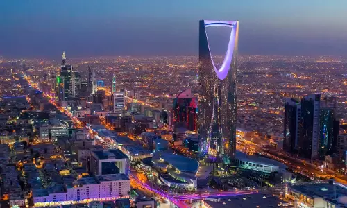 Night view of Riyadh’s skyline with the illuminated Kingdom Centre tower dominating the cityscape, surrounded by high-rise buildings and busy roads.