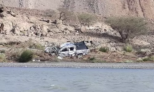 A damaged vehicle lies partially submerged near a flooded wadi in Oman, surrounded by rocky terrain after heavy rains and flash flooding.
