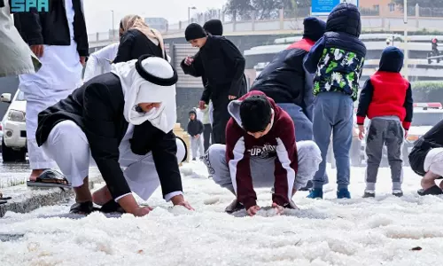 Children and adults gather outdoors in a Saudi city as hail covers the ground following a heavy storm, with people touching and examining the ice pellets.